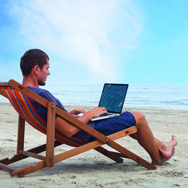 A man is on a laptop sitting by the beach.
