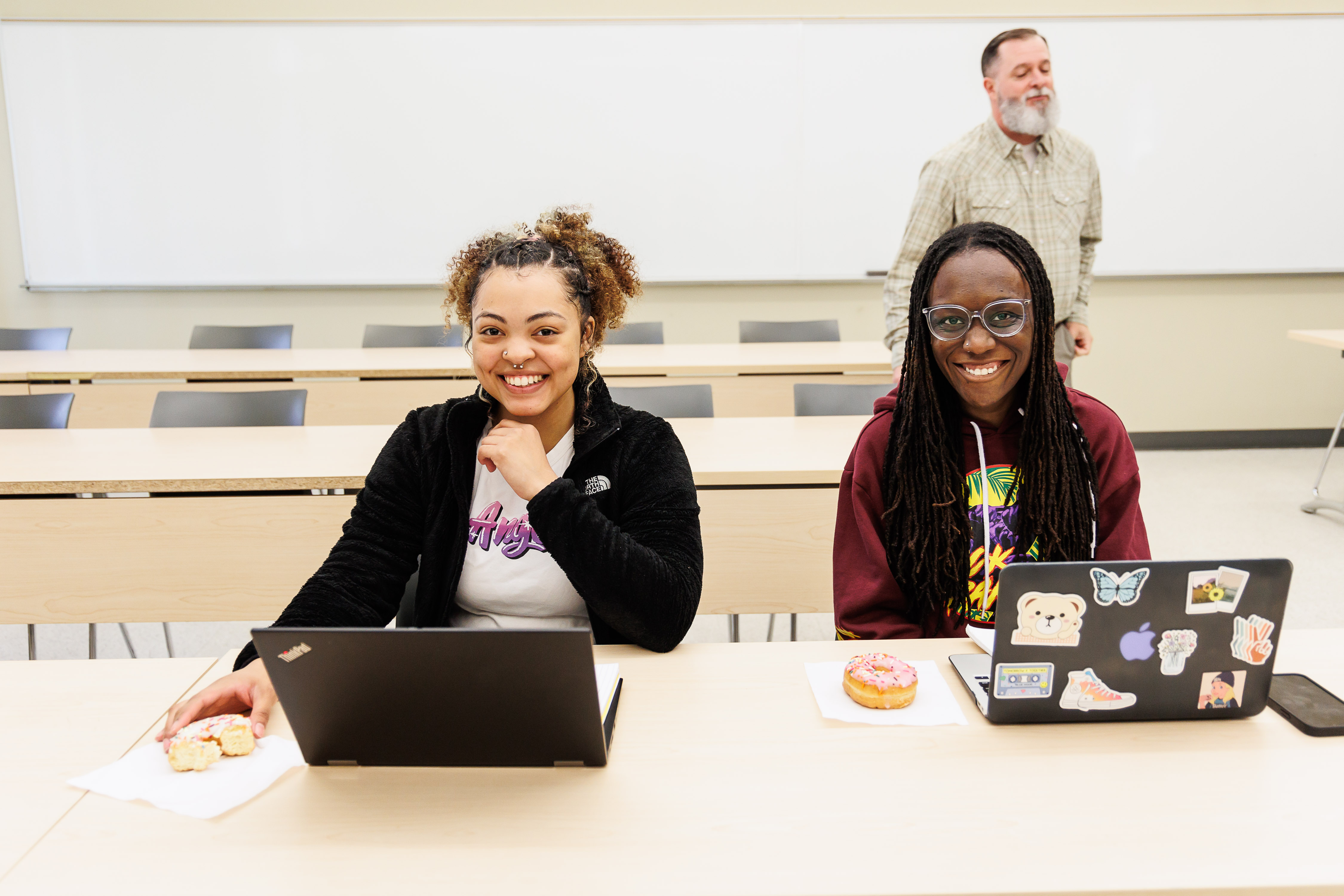 Two students are sitting in class with a snack beside their computers, smiling. 