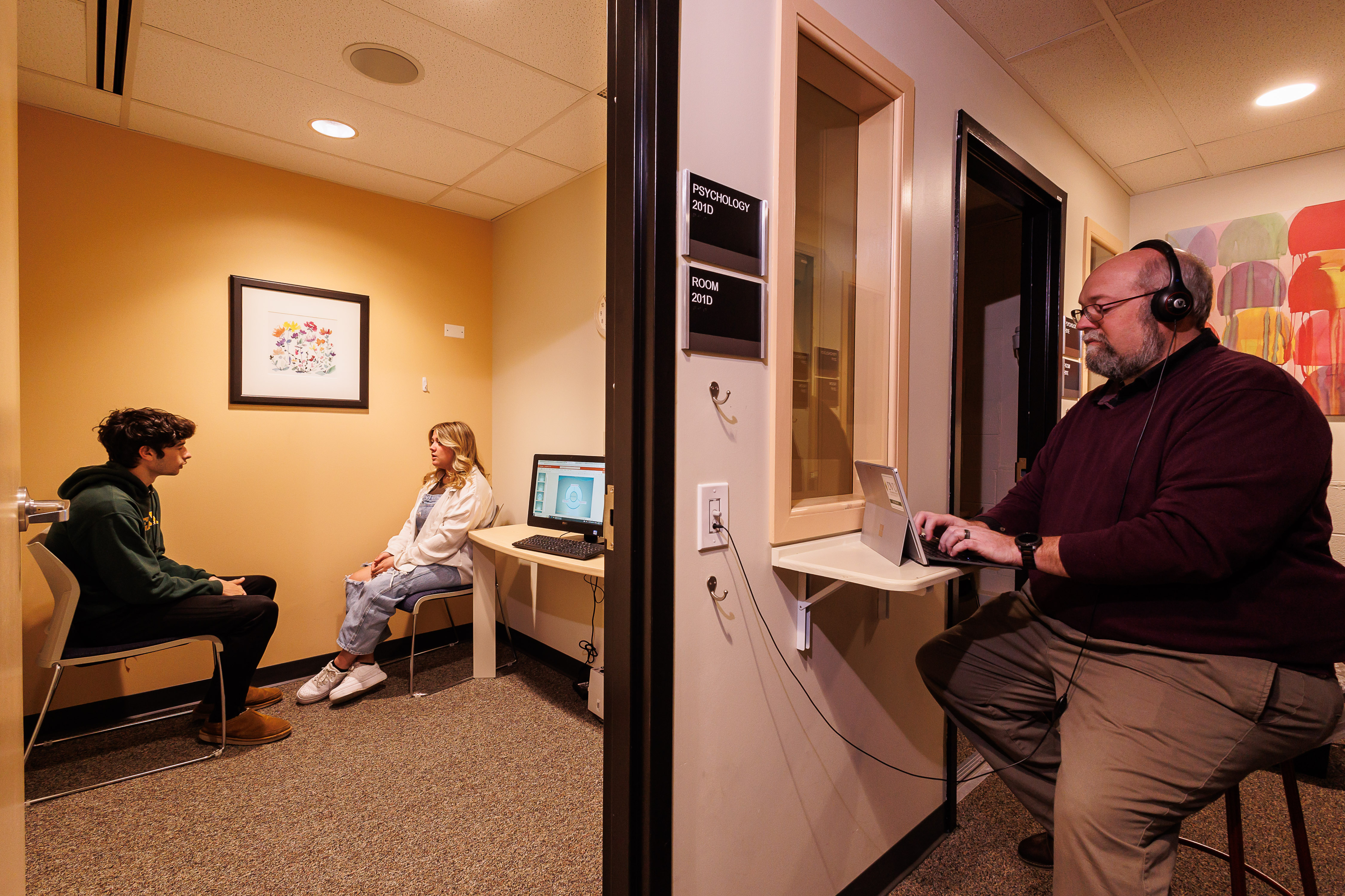 A professor is listening to a counseling session through headphones on the other side of a room with a window. This is in our counseling psychology labs. 