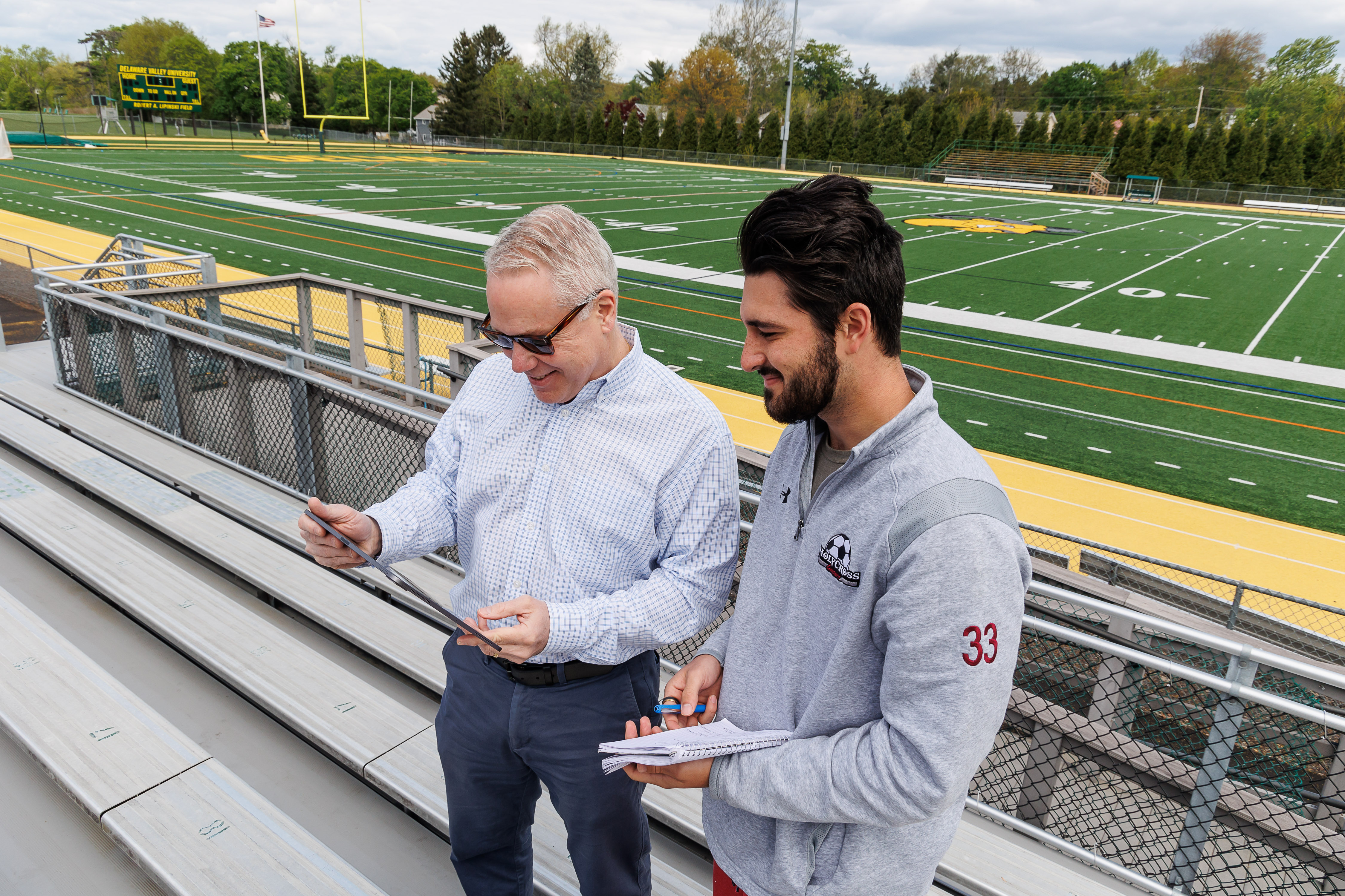 A student and professor are in the bleachers by an athletic field with notepad and tablet in hand. 