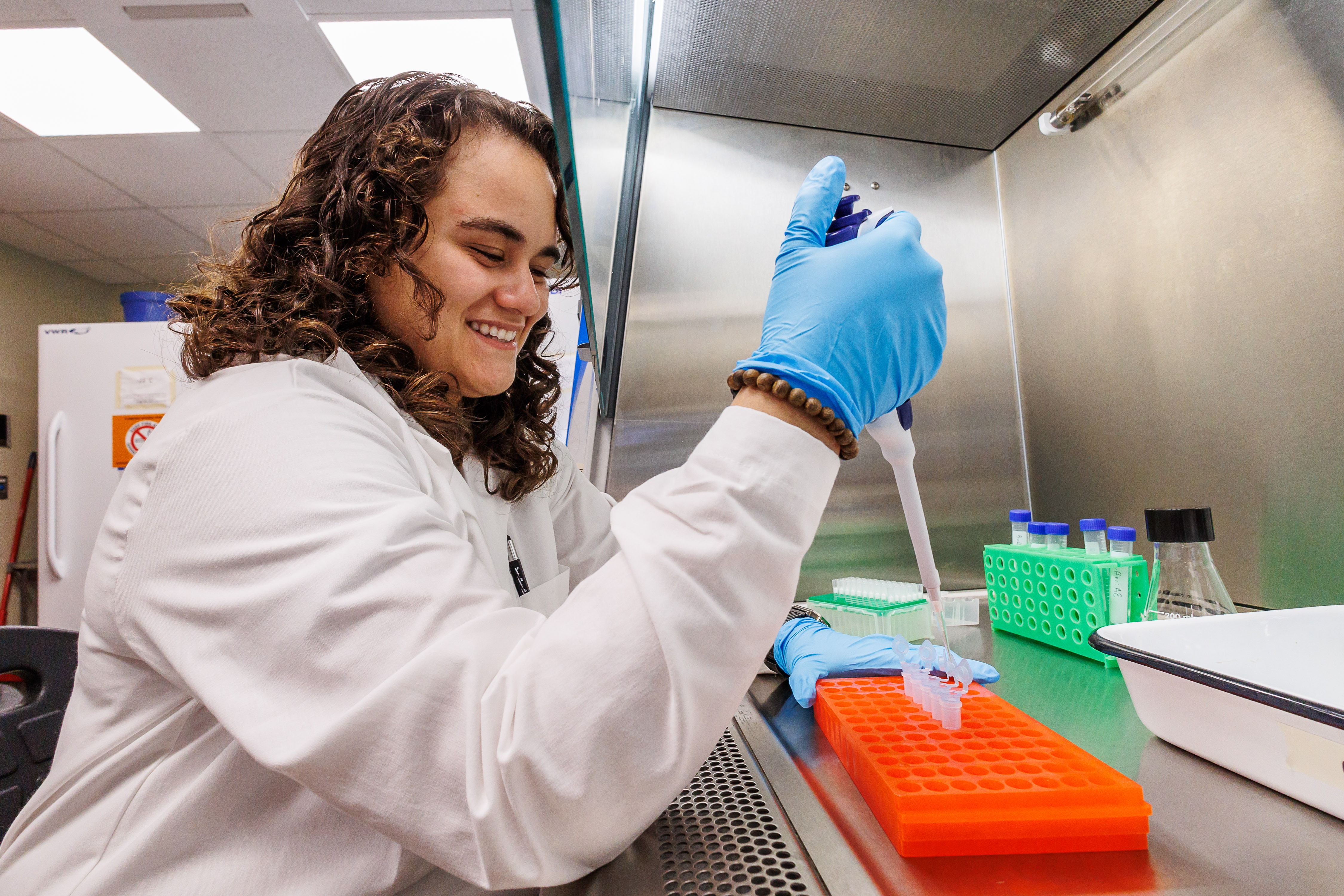 A student in a white coat and blue gloves is working in the research lab. 