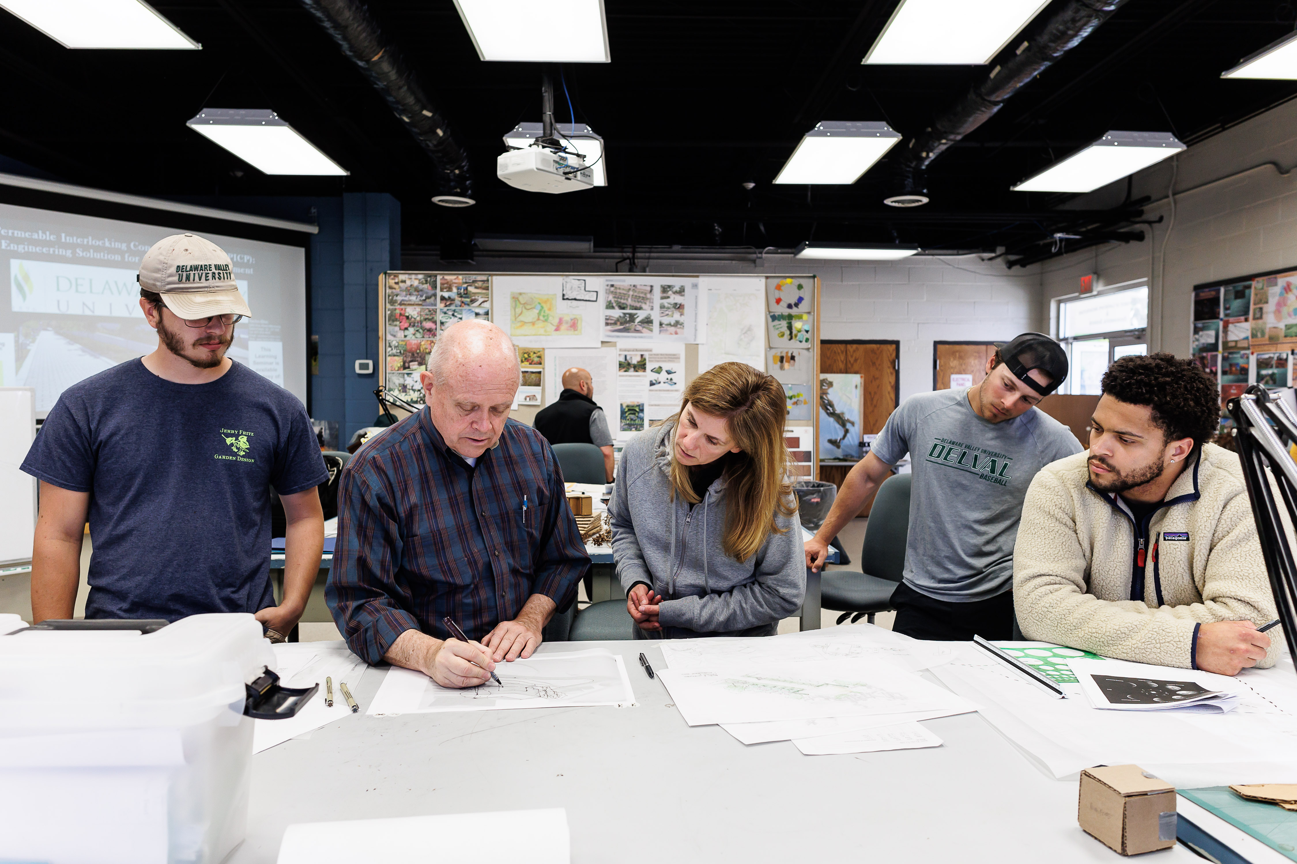 A team of professors and students are standing in front of a table with multiple hand drawn landscape designs. 