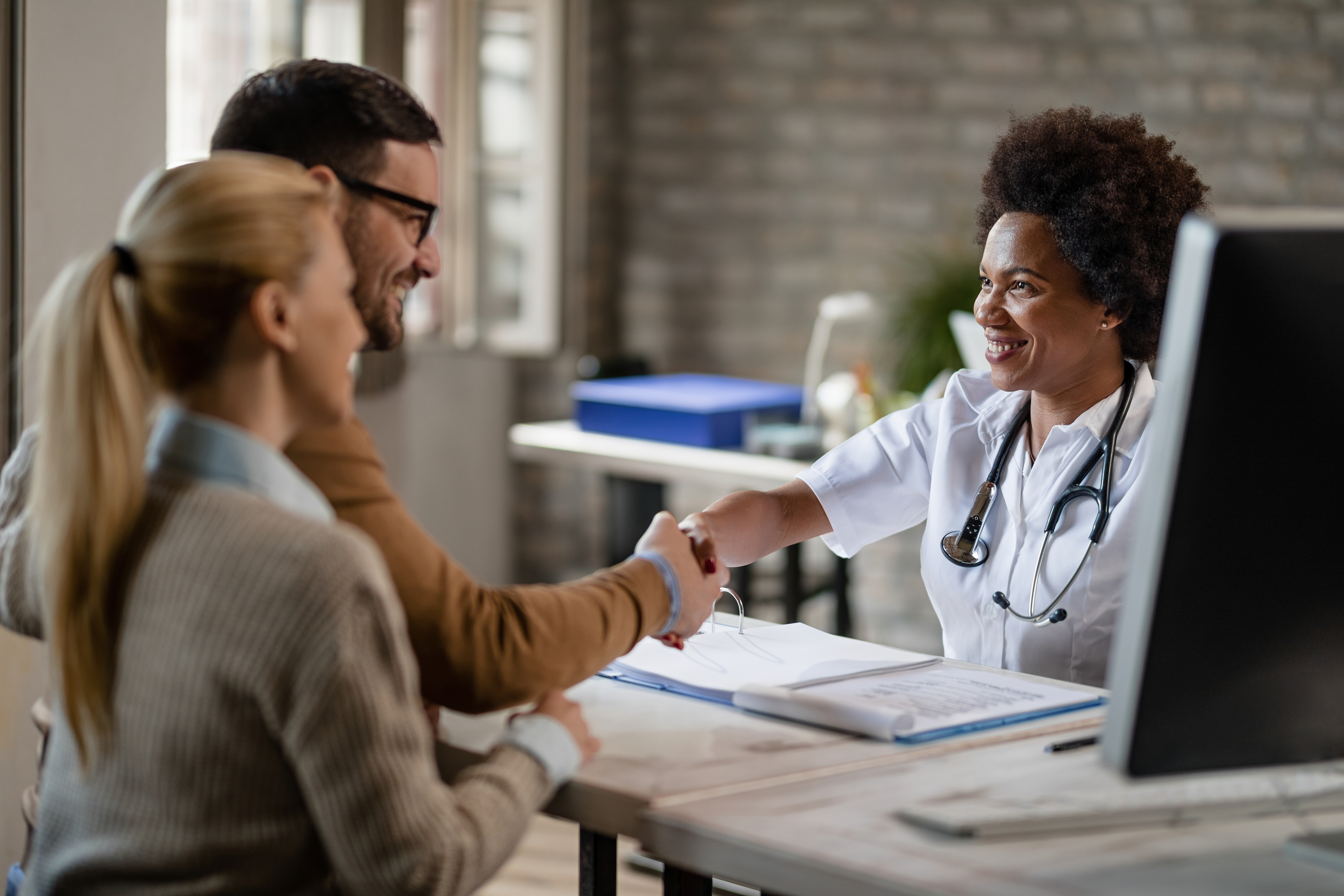A couple is shaking hands with a medical professional at a desk. 