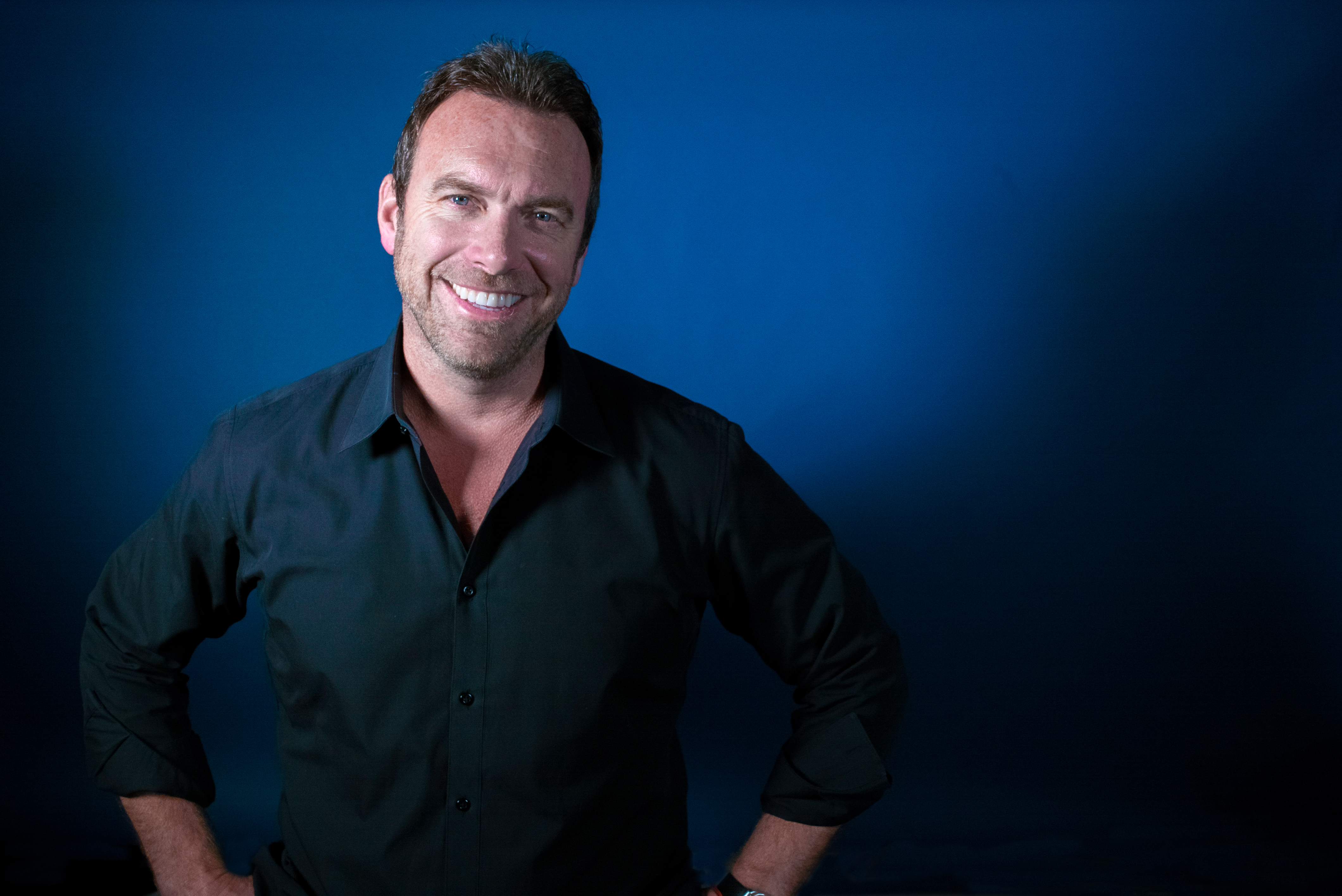 A headshot of Alex Boylan in a navy blue collared shirt in front of a royal blue background.