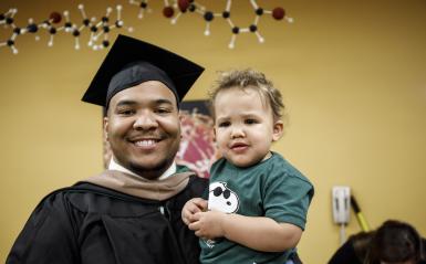 A graduate from the Continuing Studies department in his cap and gown, holding his son.