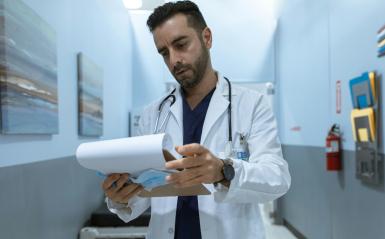 A male physician assistant is wearing a white coat and looking at a clipboard. 