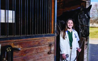 Nina Dellarte, a class of 2021 graduate is in a white coat next to a black horse at the equestrian center.