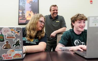 Students are working at a desk with computers, a teacher is behind them. 