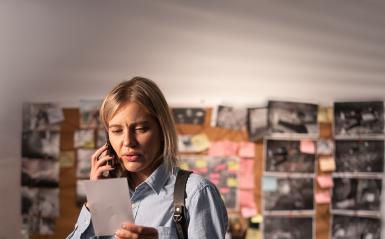 A woman is in a detective space, viewing a photo in hand while on the phone. 