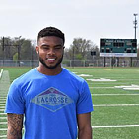 Cori Grobes stands on a field lined for football. He is wearing a Lacrosse shirt.