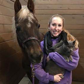 Alicia Smith, a biology alumna who completed the Honors Program at Delaware Valley University, holds a cat and stands next to a horse. 