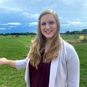 A headshot of Audrey San in a white blazer in front of a field of grass.