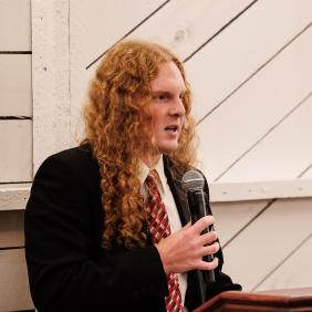 Caleb Messenger is at a podium speaking into a microphone at an event on campus.