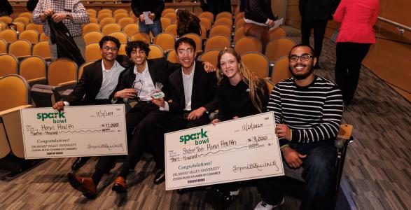 Katia Holmes and Cory Korkins, who students of DelVal, sit with the Spark Bowl winning business while holding giant checks. 