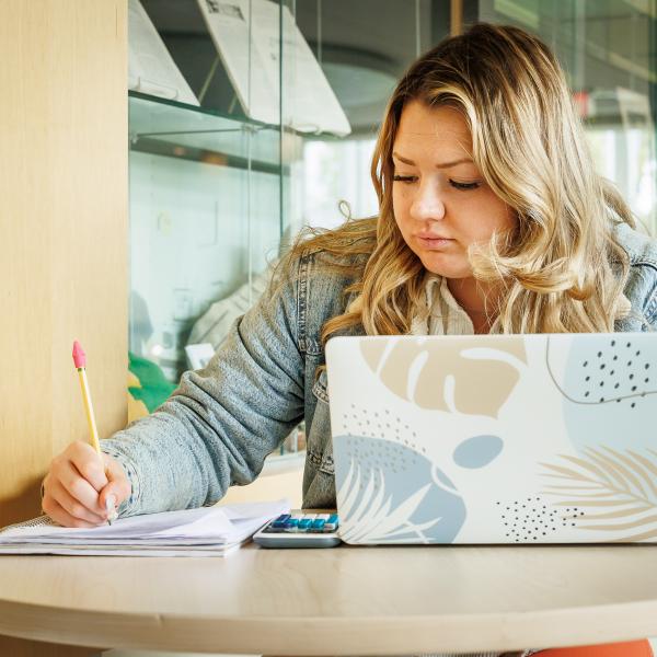 A student is writing in a note book while her laptop sits open on the table in front of her. 