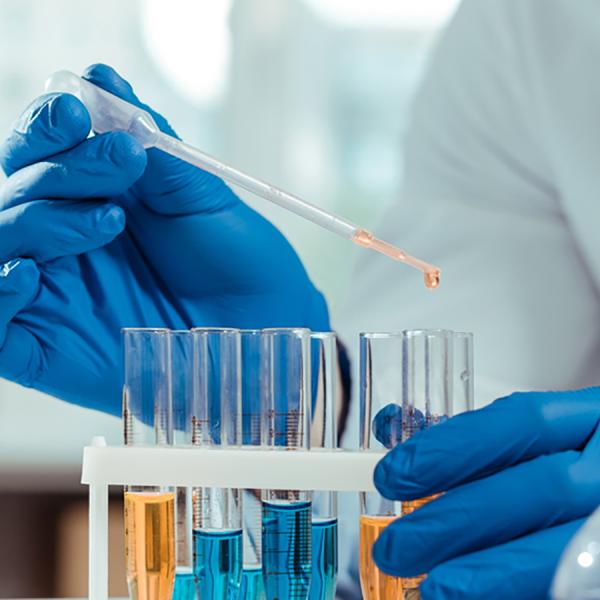 Someone wearing blue gloves is dropping liquid into a beaker in a chemistry lab, the view is of the hands only.