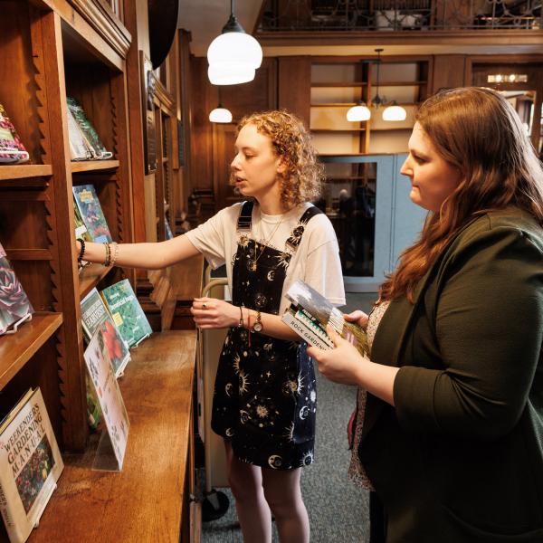 A student is in the library looking at books and speaking to another staff member. 