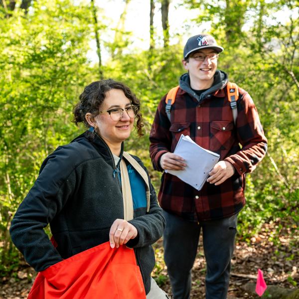 Two students are exploring outdoors in a wooded area with backpacks.