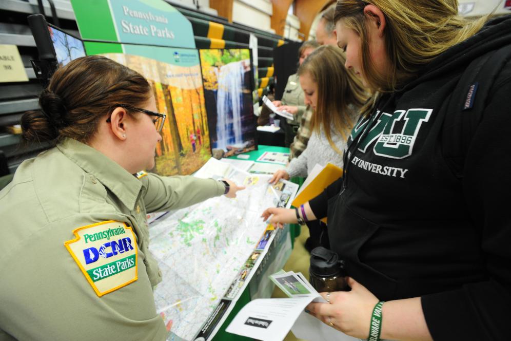 A uniformed member of the Pennsylvania Department of Conservation and Natural Resources points toward an information table while speaking with two Delaware Valley University students about job opportunities in conservation.