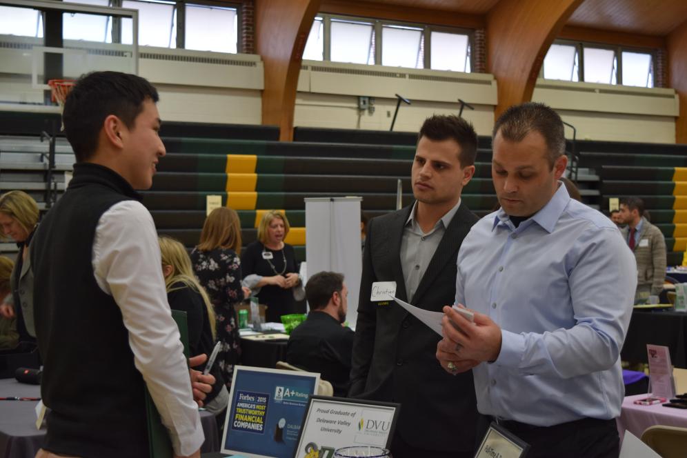 Two employer representatives stand behind an information table and review paperwork while speaking with a Delaware Valley University student about job opportunities at the career and internship fair. 