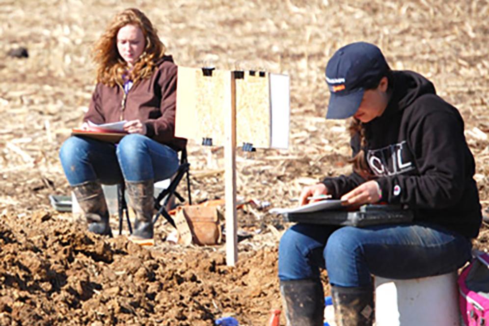 Two students sitting on portable chairs taking soil samples in a field.