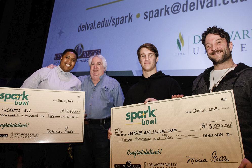 Four people standing on a stage with holding two prize money checks won from DVU's Spark Bowl business competition.