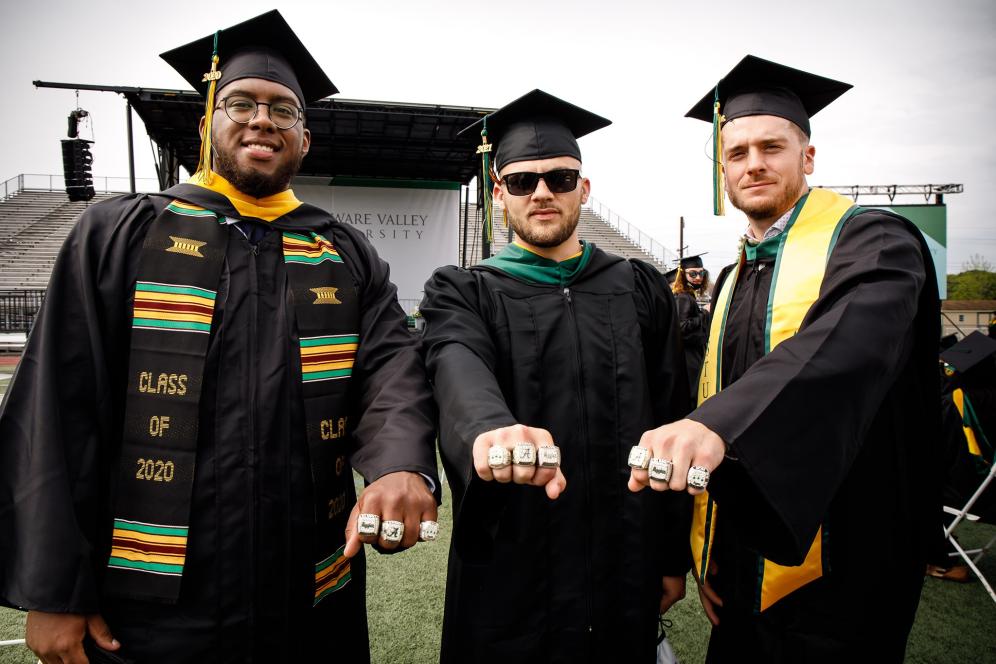 Three graduates, in their cap and gowns, showing their class rings.