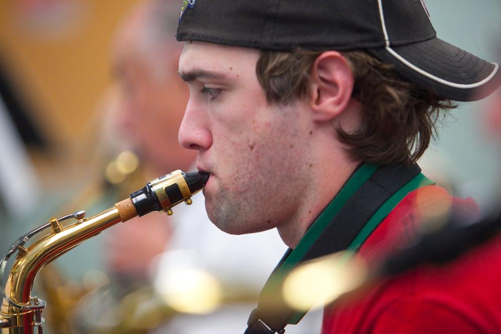A sitting student wearing a black hat and red shirt is blowing into a tuba.