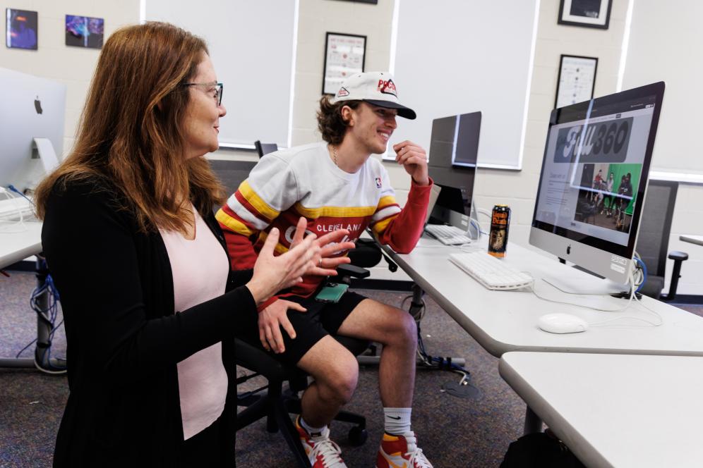 Marion is working with a male student indoors in front of a mac computer. 