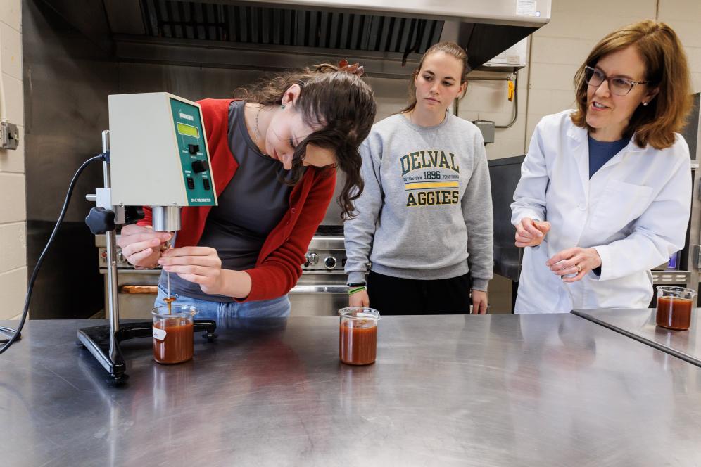 Laura Herbst is in the food lab with two students using food science technology to measure liquid in beakers.