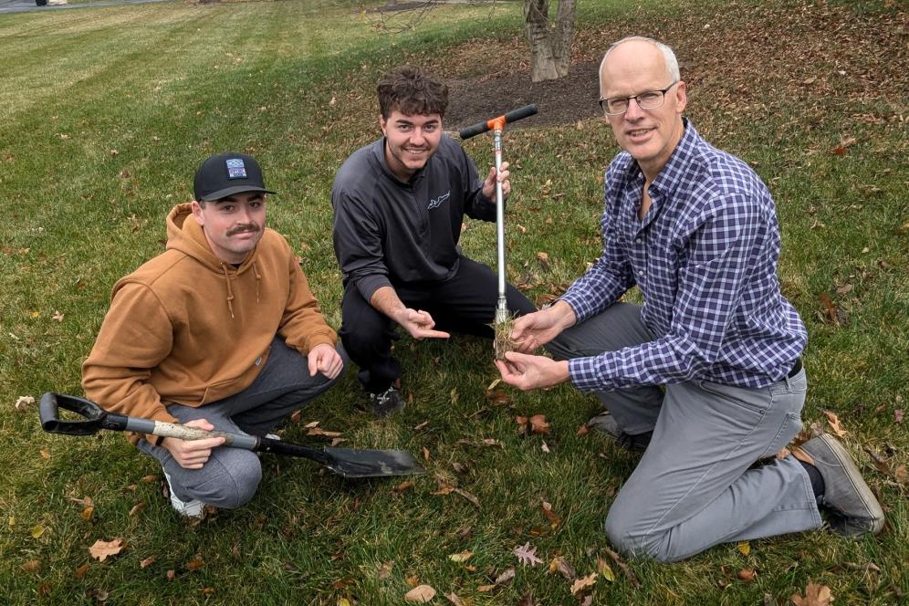 Two students are on the ground working with Dr. Linde in a grassy field.