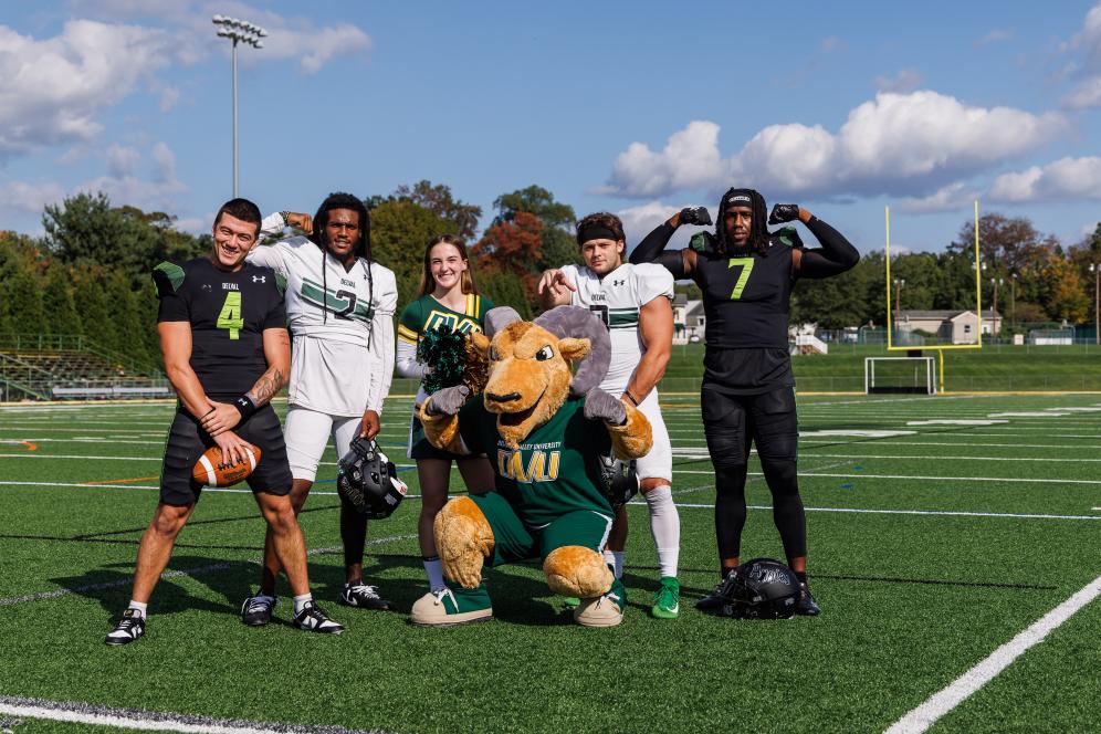 Students from the Football team and one cheerleader are posing on the field with Caesar the ram mascot.