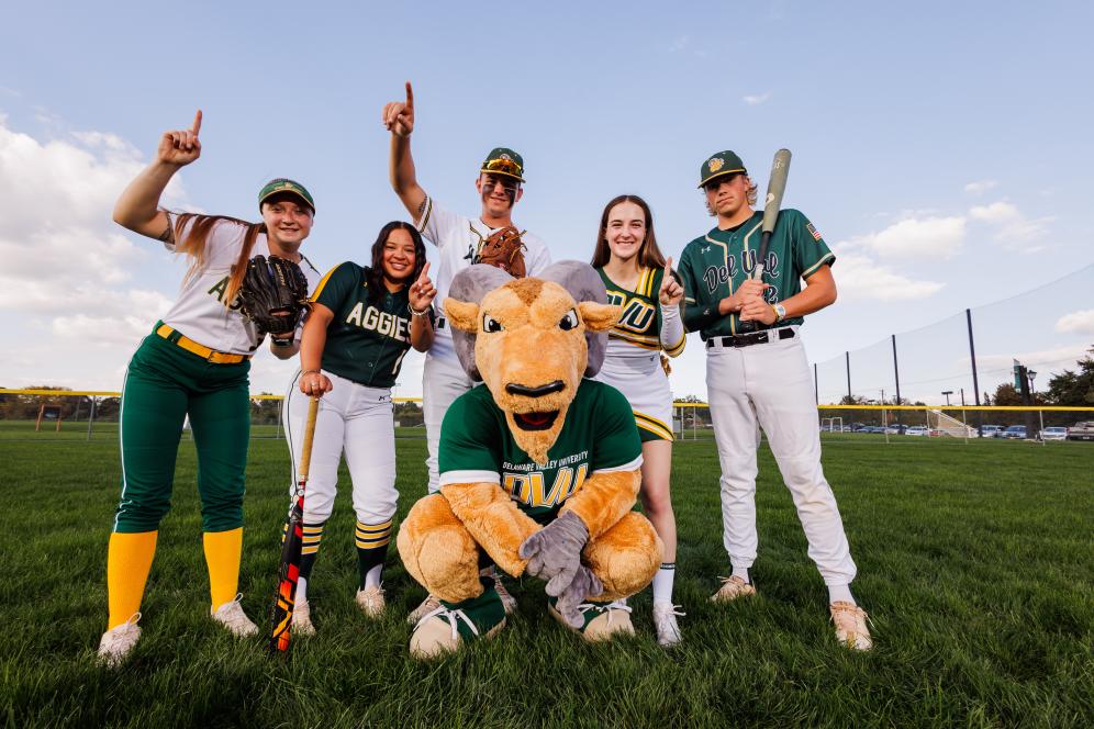 Students from the baseball team and softball team and one cheerleader are posing on the field with Caesar the ram mascot.
