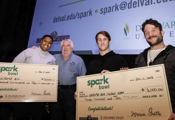 Four people standing on a stage with holding two prize money checks won from DVU's Spark Bowl business competition.