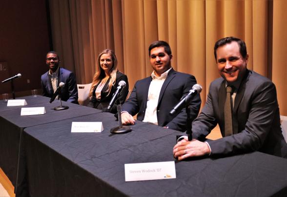 Four speakers are seated on stage in formal business attire. 