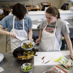 male and female students wearing aprons and plating food from a large frying pan