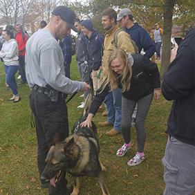 A police officer and a K9 dog are standing in front of a female student who is petting the dog.