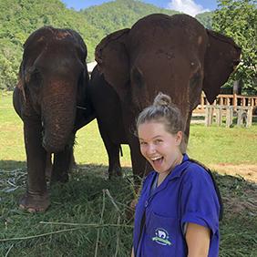 College-age student standing in from of 2 elephants at a sanctuary