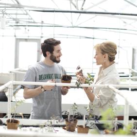 A student and faculty member are in the greenhouse talking about plants. 