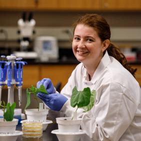 A student in the lab with a potted plant in gloves and white coat. 