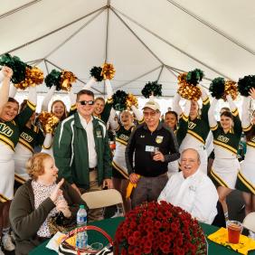 Dr. Ben is with students and alumni at homecoming celebrating. Cheerleaders are yelling in the background.