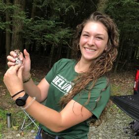 Alumna Chrissy Lambert is holding a small animal in a wooded area.