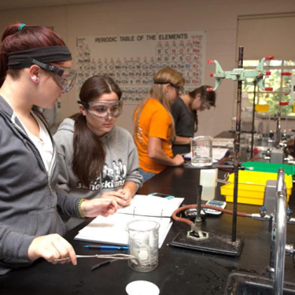 Students at a lab table taking notes in front of chemistry equipment