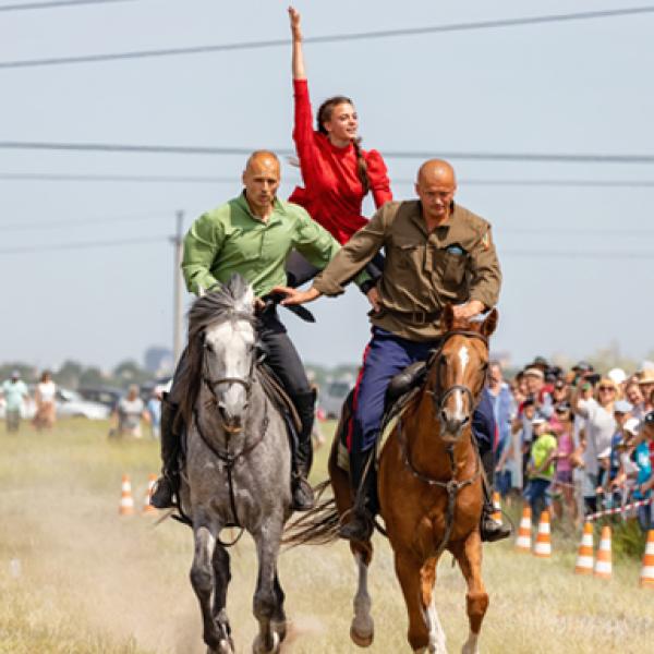 Two male students ride horses. A female student stands with a leg on either horse in an acrobatic pose.