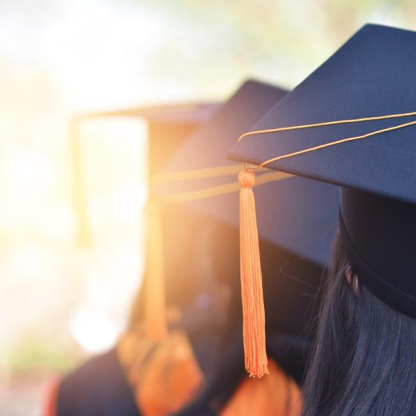 Graduates sit in file. We see their caps.