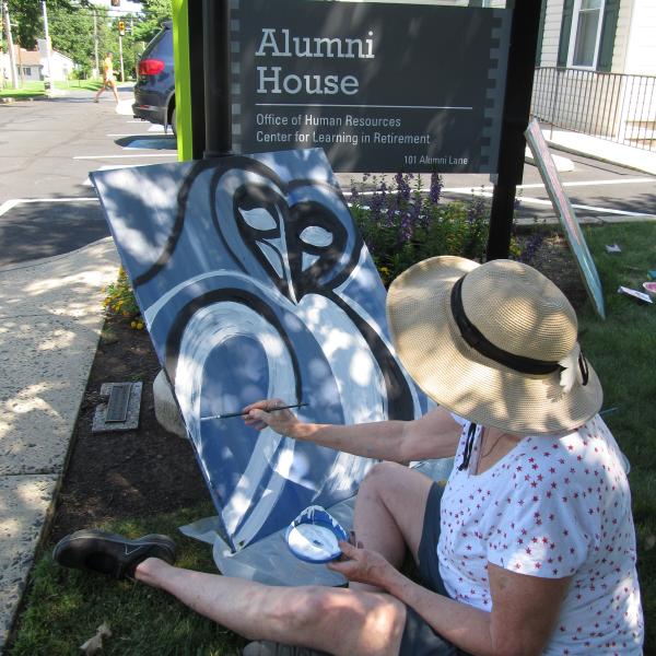 An older woman sitting on the ground painting an owl on a 18x24 canvas