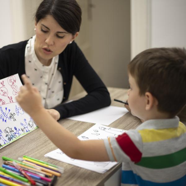 A woman is working with a child at a table with paper and crayons. 