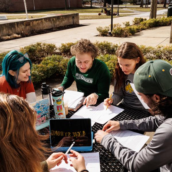 Students are outdoors with. their laptops studying on a bench.