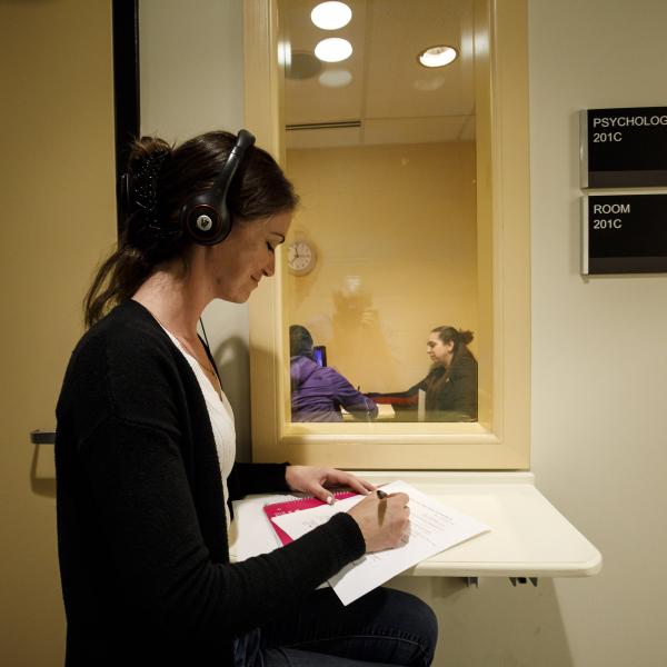 A counseling psychology student is listening to a session through earphones from outside of a room while taking notes. 