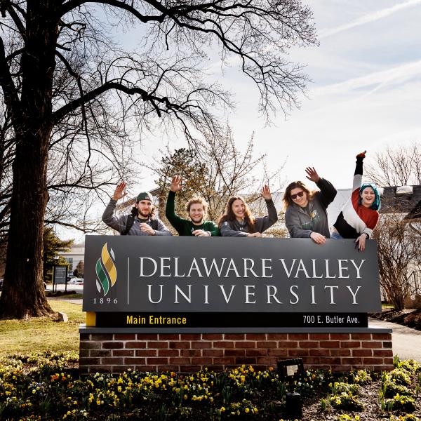 A few students are waiving at the camera standing behind a large delval sign.